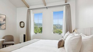 A hotel room with a large bed, two windows with curtains, a view of the city, a round mirror, and a chair. - Casa Riviera 24 - Boutique Suites (Santa Barbara)