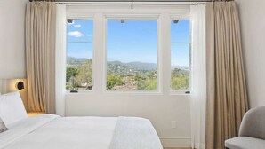 Una camera da letto con un letto grande, due finestre con tende e vista sulla città, più una sedia.