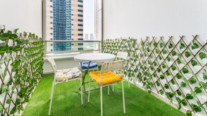 Un balcon avec une table et des chaises blanches, un gazon artificiel vert et une vue sur de hauts immeubles.