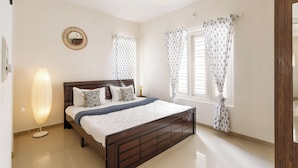 A bedroom with a wooden bed, a ceiling fan, a bedside lamp, and patterned curtains. - Aira Luxe by Red Olivebyg Brewskiwifi (Bengaluru)