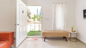 A room with a bed, a bench, a small table, and a door leading to an outdoor area with plants. - Aira Luxe by Red Olivebyg Brewskiwifi (Bengaluru)