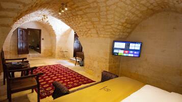 Una habitación con techo de piedra, una cama, un televisor montado en la pared, una silla de madera y una alfombra roja estampada.