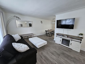 A modern living room with a black sofa, white ottoman, dining table with chairs, a TV on a stand, and a fireplace.