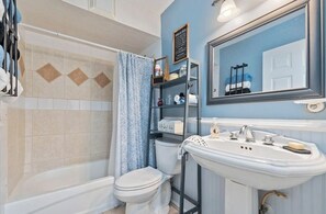 A bathroom with a bathtub, toilet, and sink. There is a shower curtain, a shelf with toiletries, and a mirror above the sink.