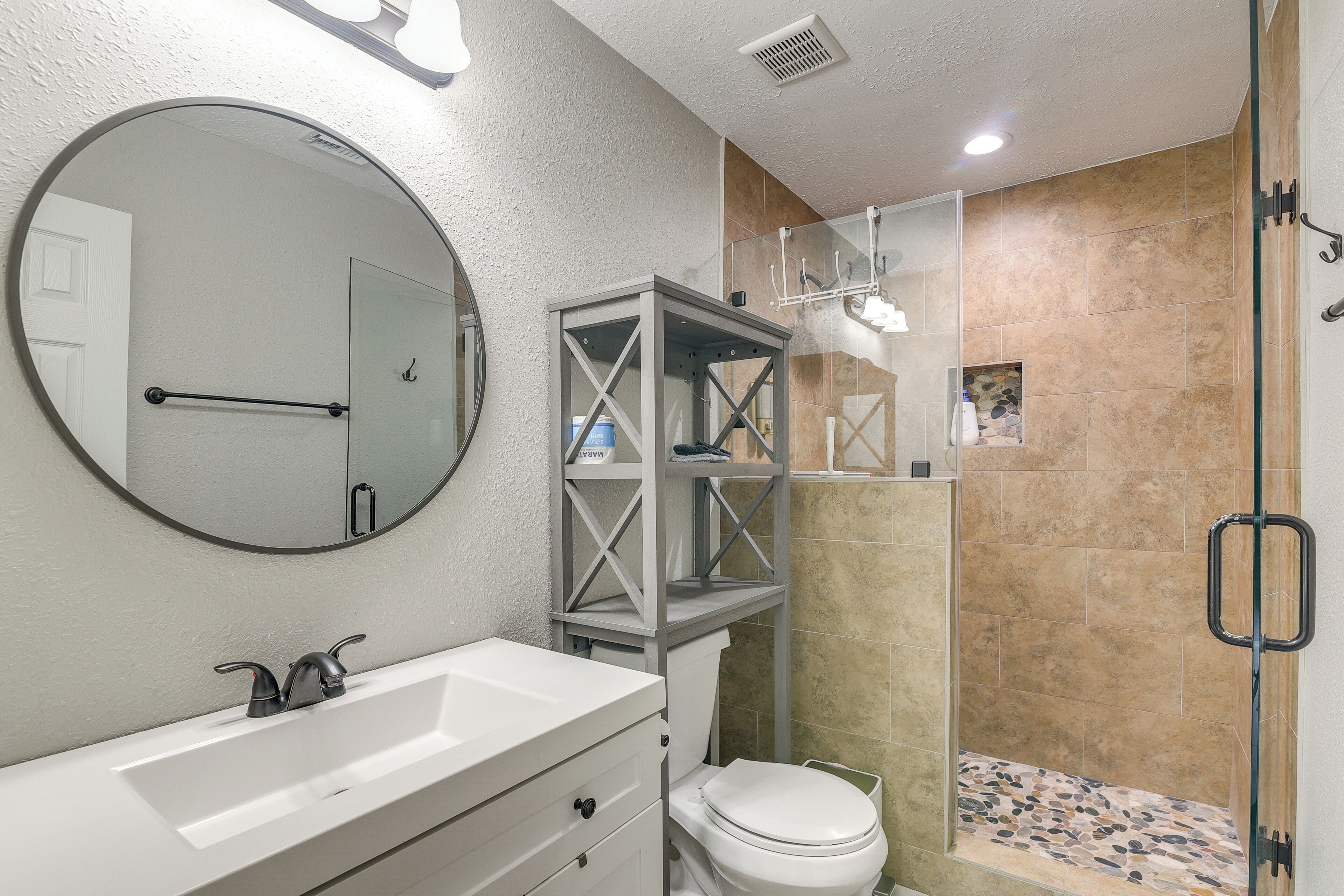 Une salle de bain avec un lavabo blanc, un miroir rond, une douche à parois de verre et des toilettes.