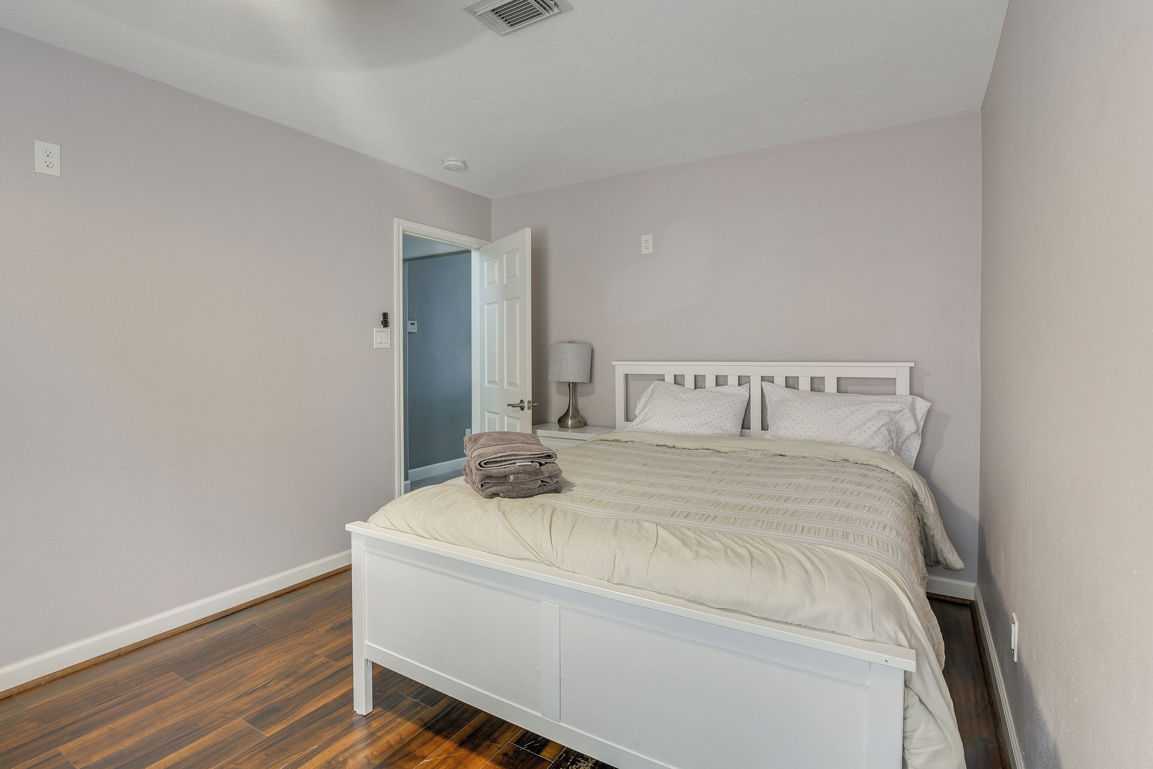 Un dormitorio con una cama blanca, una puerta que da a otra habitación y una lámpara sobre una mesita de noche.