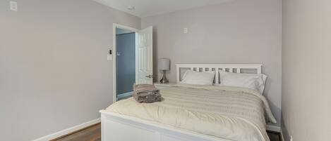 Un dormitorio con una cama blanca, una puerta que da a otra habitación y una lámpara sobre una mesita de noche.