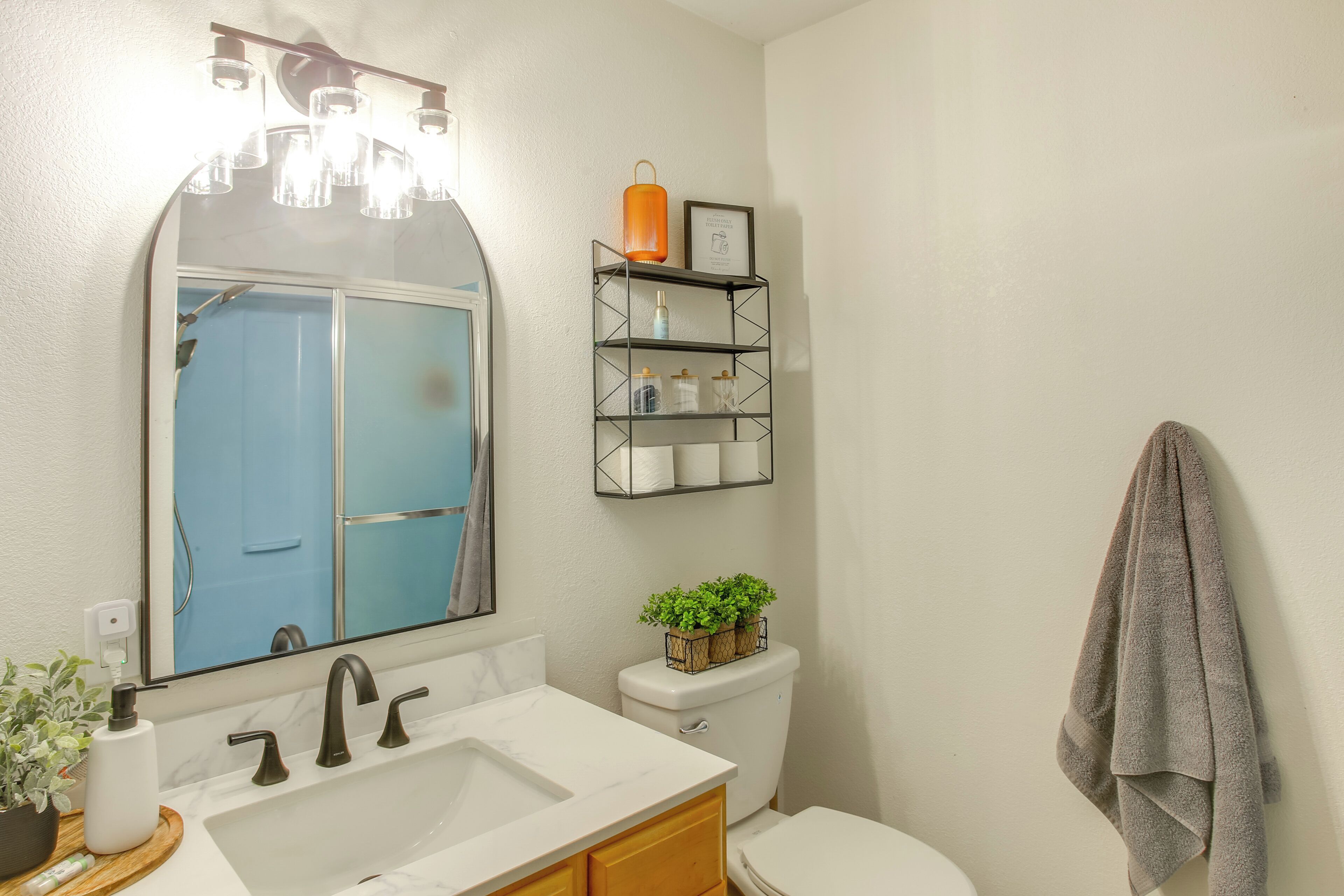 Un baño con lavamanos, espejo, inodoro y estantes con artículos de aseo.