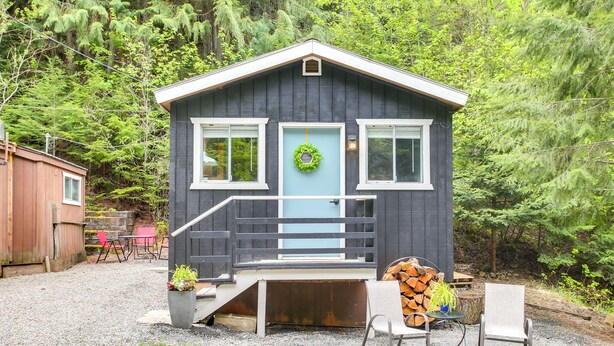 A small cabin with a front porch, a set of stairs, and a door with a wreath. There are chairs and a table outside, and a stack of firewood nearby. - Hike & Fish: Mountain Getaway in Avery! (Avery)