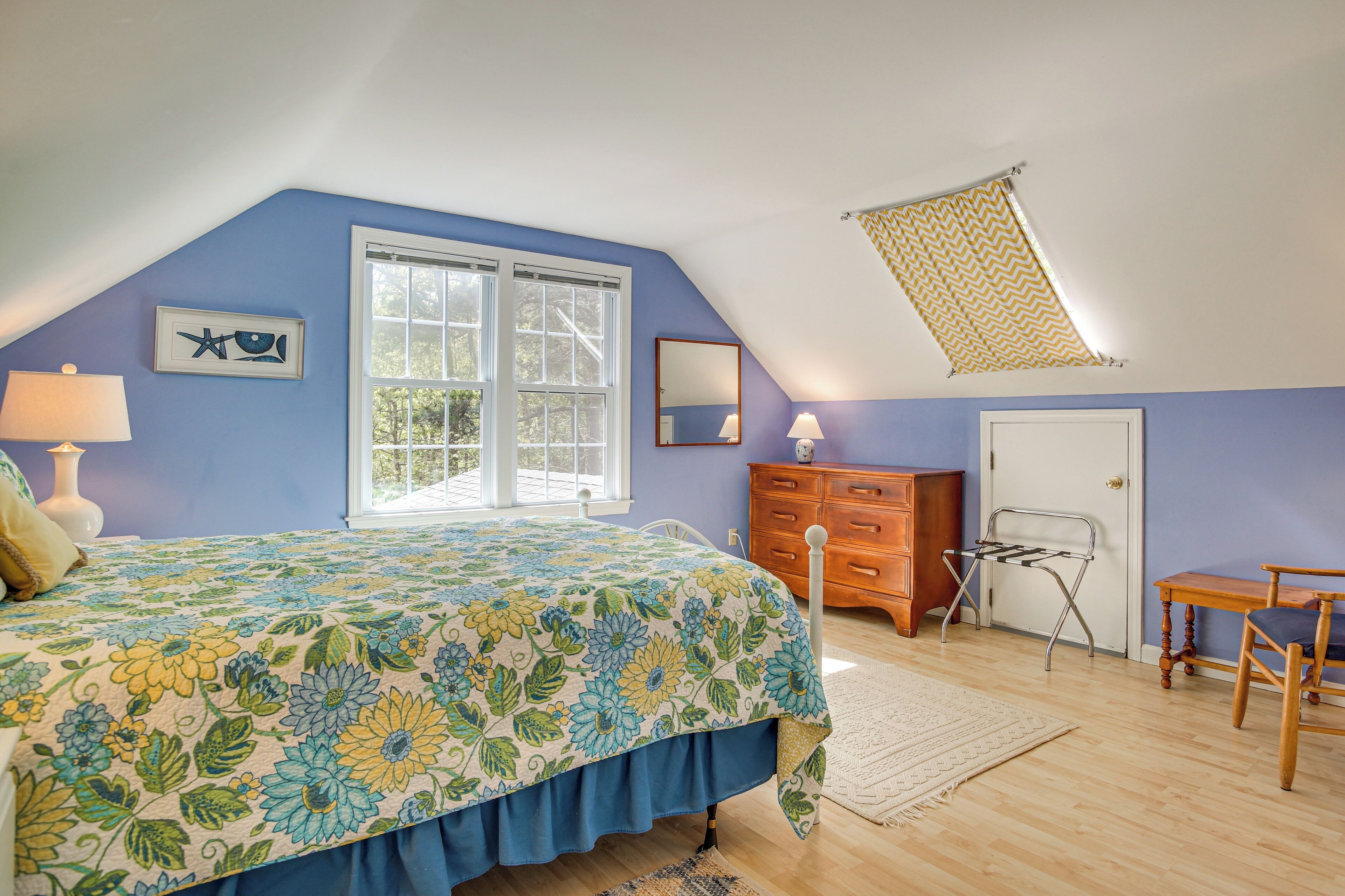 Ein Schlafzimmer mit schrägen Wänden, einem Bett mit blumigem Überwurf, einem Schminktisch, einem Stuhl und einem Fenster mit Blick auf Bäume.