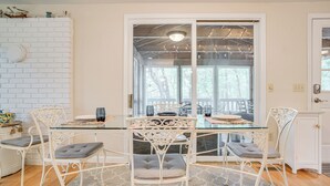 A dining area with a glass table and white metal chairs, a white brick wall, and a view through a glass door to an outdoor deck. - 1/2 Mi to Cooks Brook Beach: Cape Cod Gem w/ Deck (Eastham)