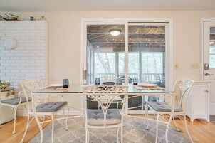 Un comedor con una mesa de vidrio y sillas de metal blanco, una pared de ladrillo blanco y vistas a una terraza exterior a través de una puerta de cristal.