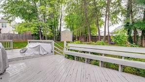 Une terrasse en bois avec un barbecue couvert, un banc et un abri de jardin, le tout situé dans une cour arrière bordée d’arbres.