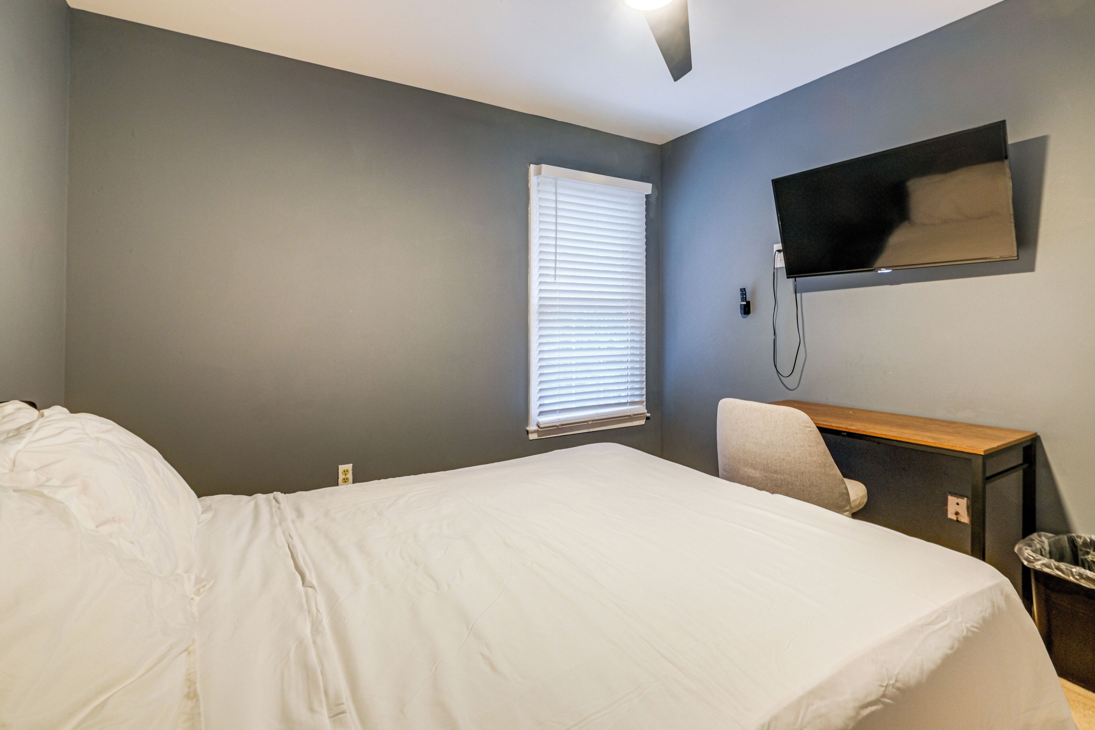A bedroom with a bed, a desk, a chair, a TV mounted on the wall, and a window with blinds.