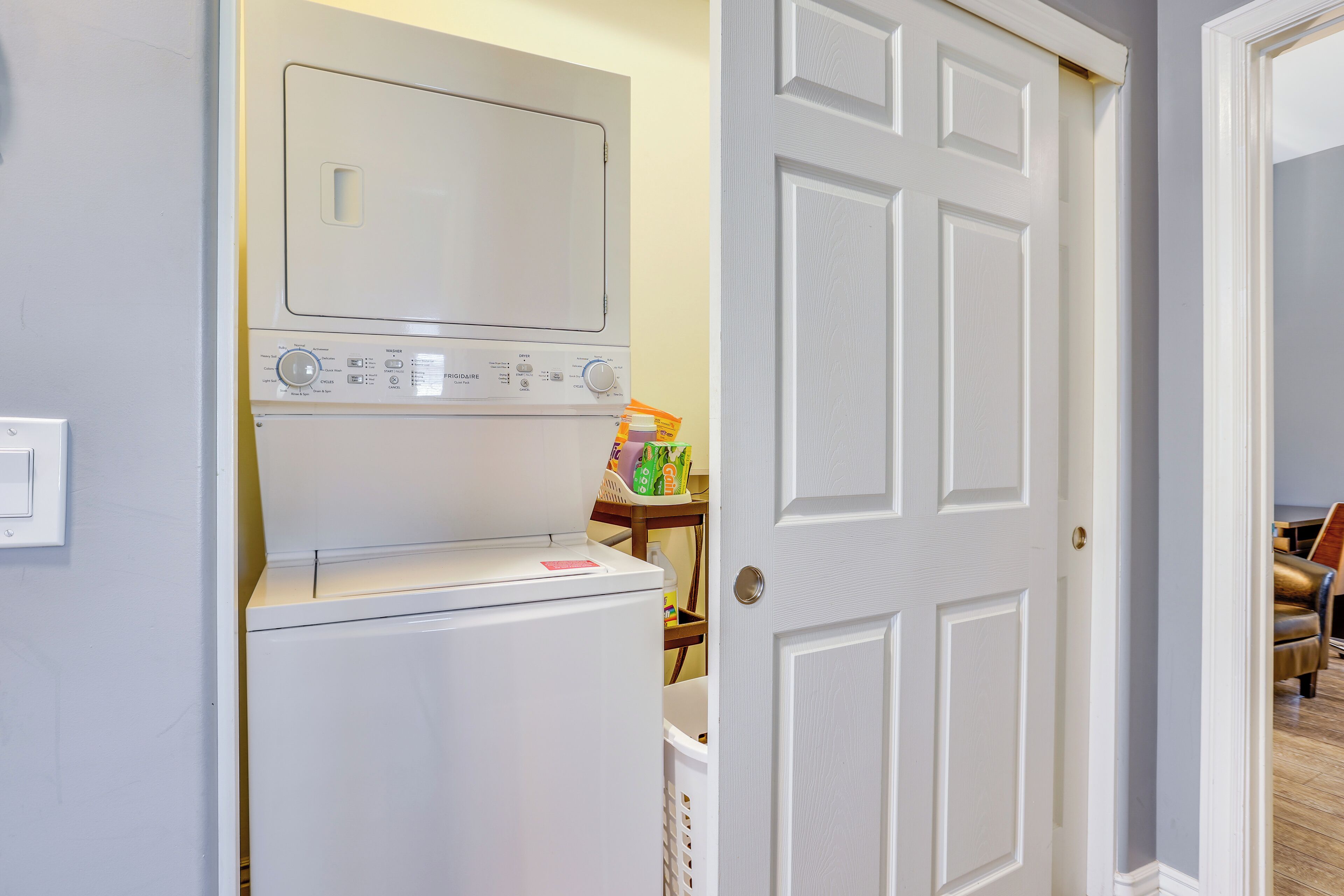 Zona de lavandería con lavadora y secadora, una mesita con productos de limpieza y una puerta que da a otra habitación.