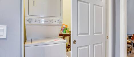 Zona de lavandería con lavadora y secadora, una mesita con productos de limpieza y una puerta que da a otra habitación.