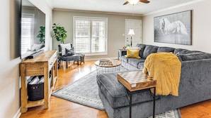 A living room with a gray sectional sofa, a wooden coffee table, a TV on a wooden stand, a ceiling fan, and a large window with blinds. - Covered Patio! Modern Home in Jackson (Jackson)
