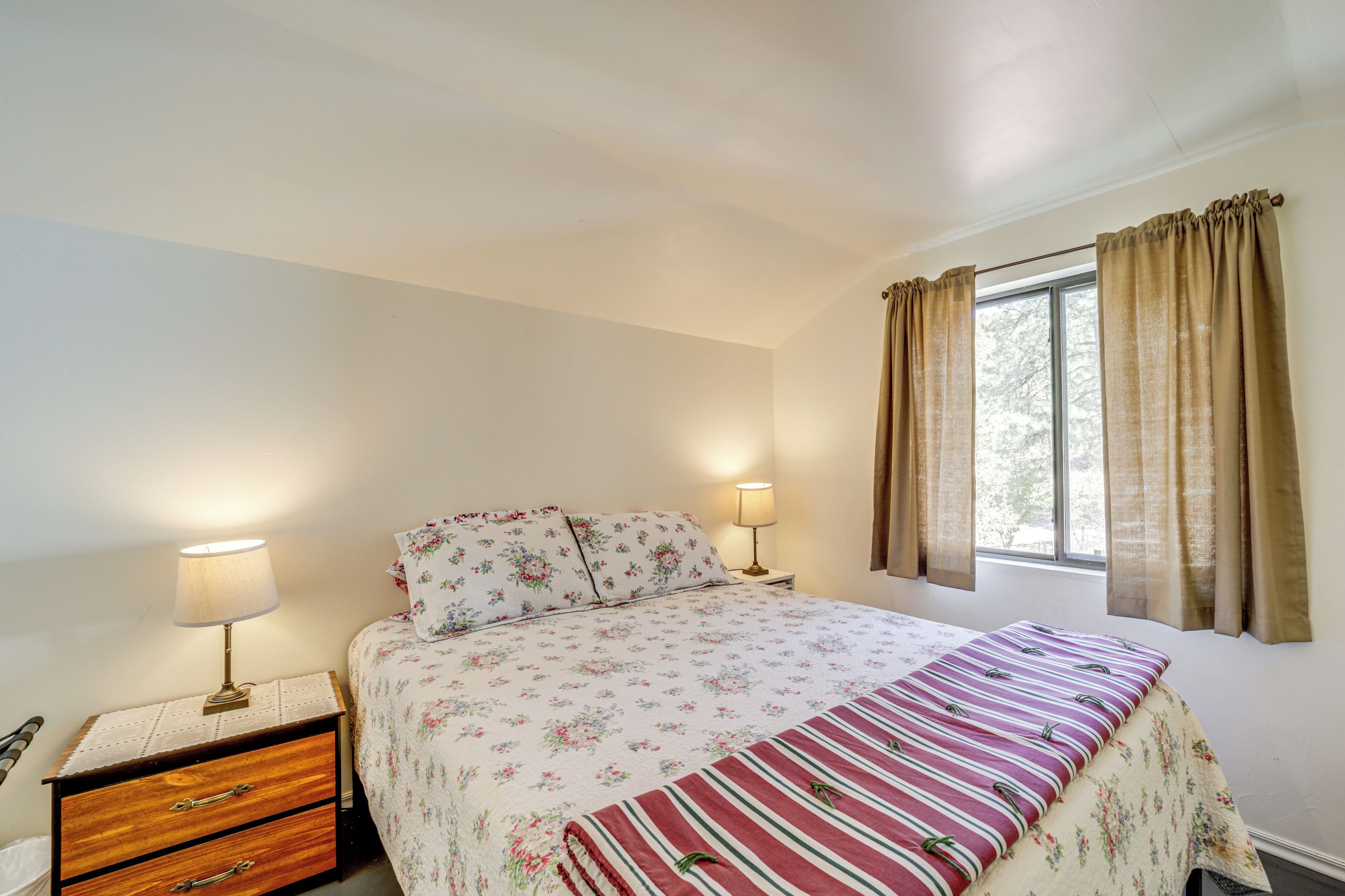 Une chambre à coucher avec un lit, deux lampes de chevet, une fenêtre avec des rideaux et une table de chevet en bois dotée de tiroirs.