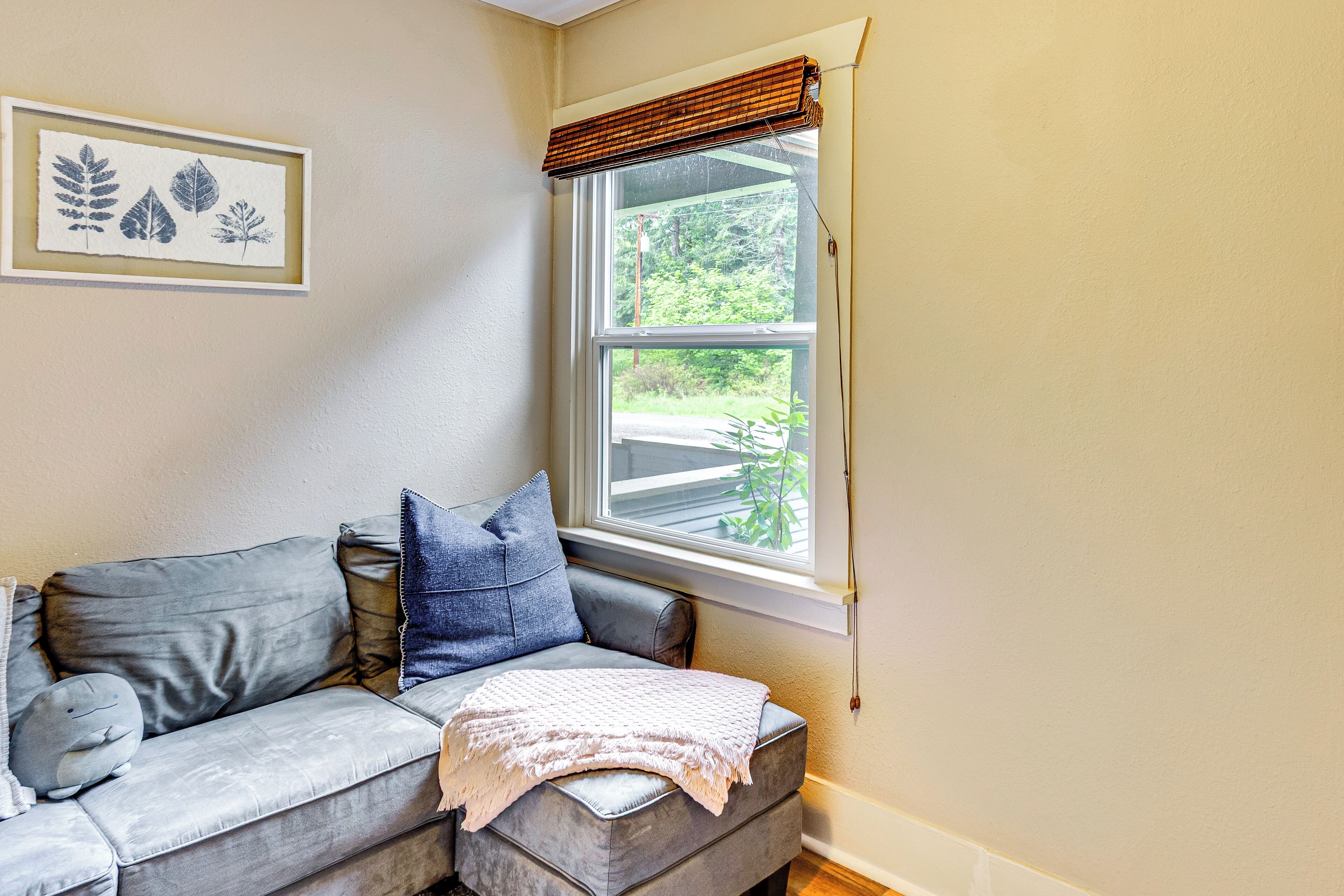 A corner room with a grey sofa, a framed botanical print on the wall, and a window with a blind.