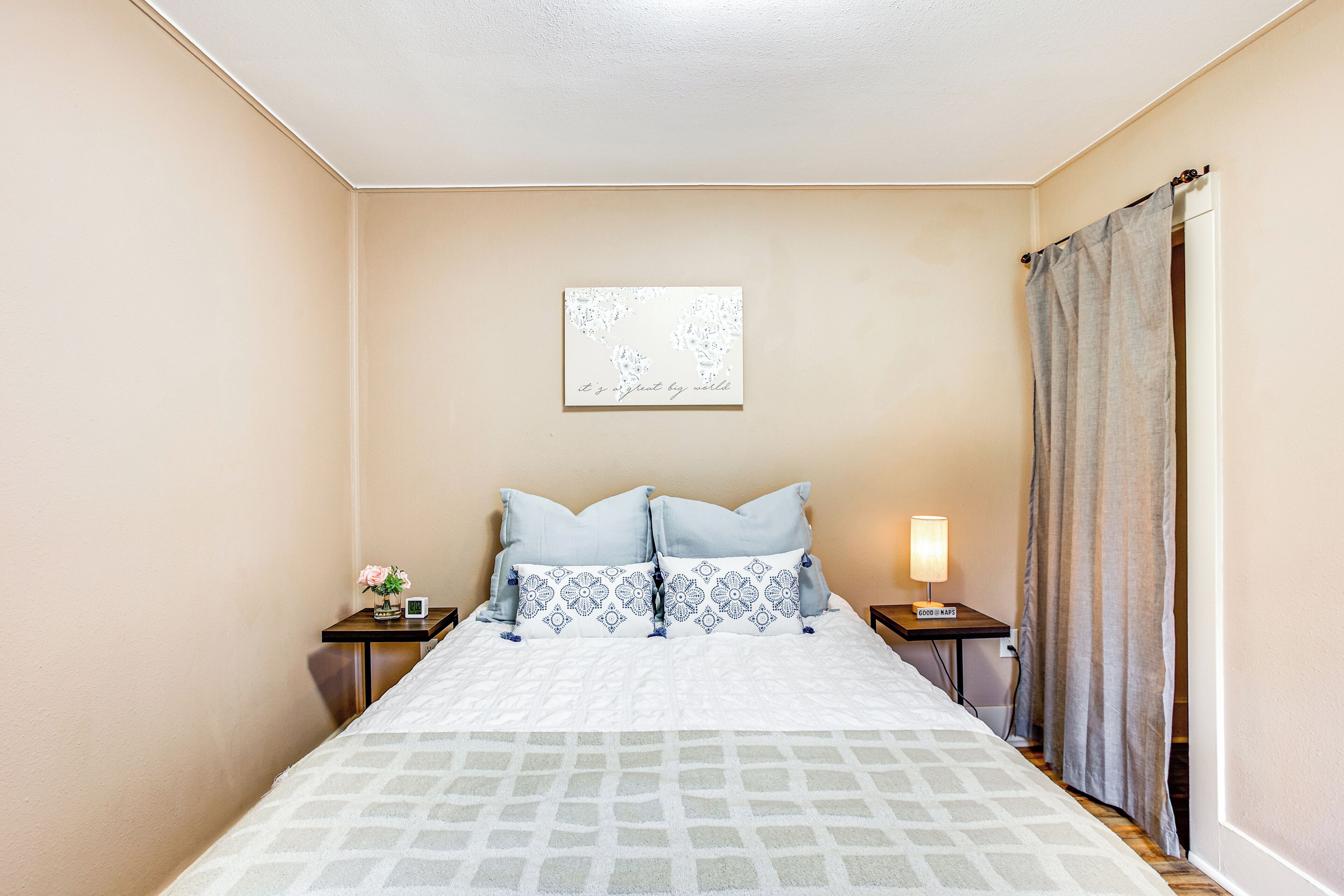 A neatly arranged bedroom with a bed, two bedside tables, a nightstand, a wall-mounted artwork, and a curtain.
