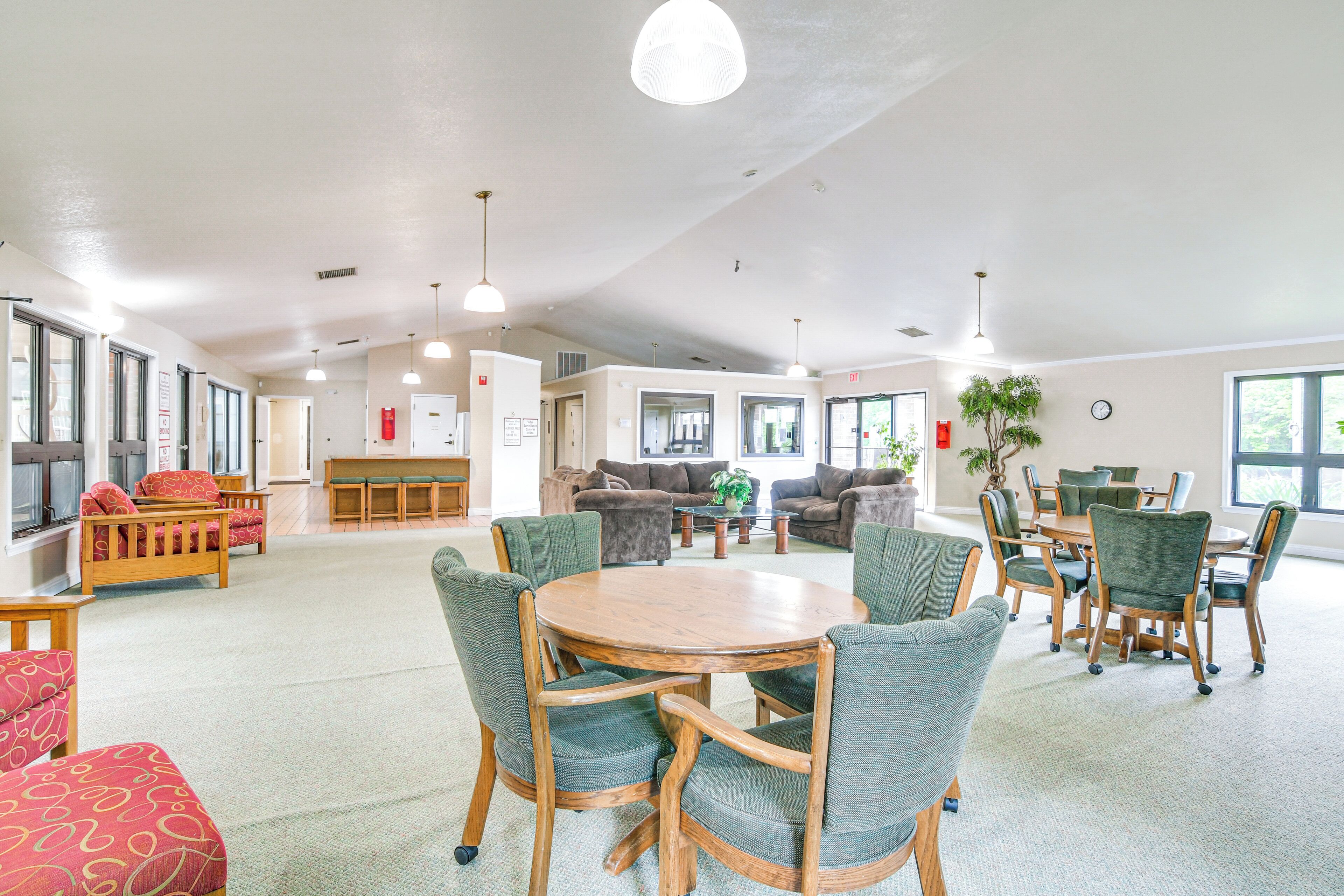 A spacious room with a round wooden table surrounded by green upholstered chairs, a sofa set, and a dining area with a wooden table and chairs.