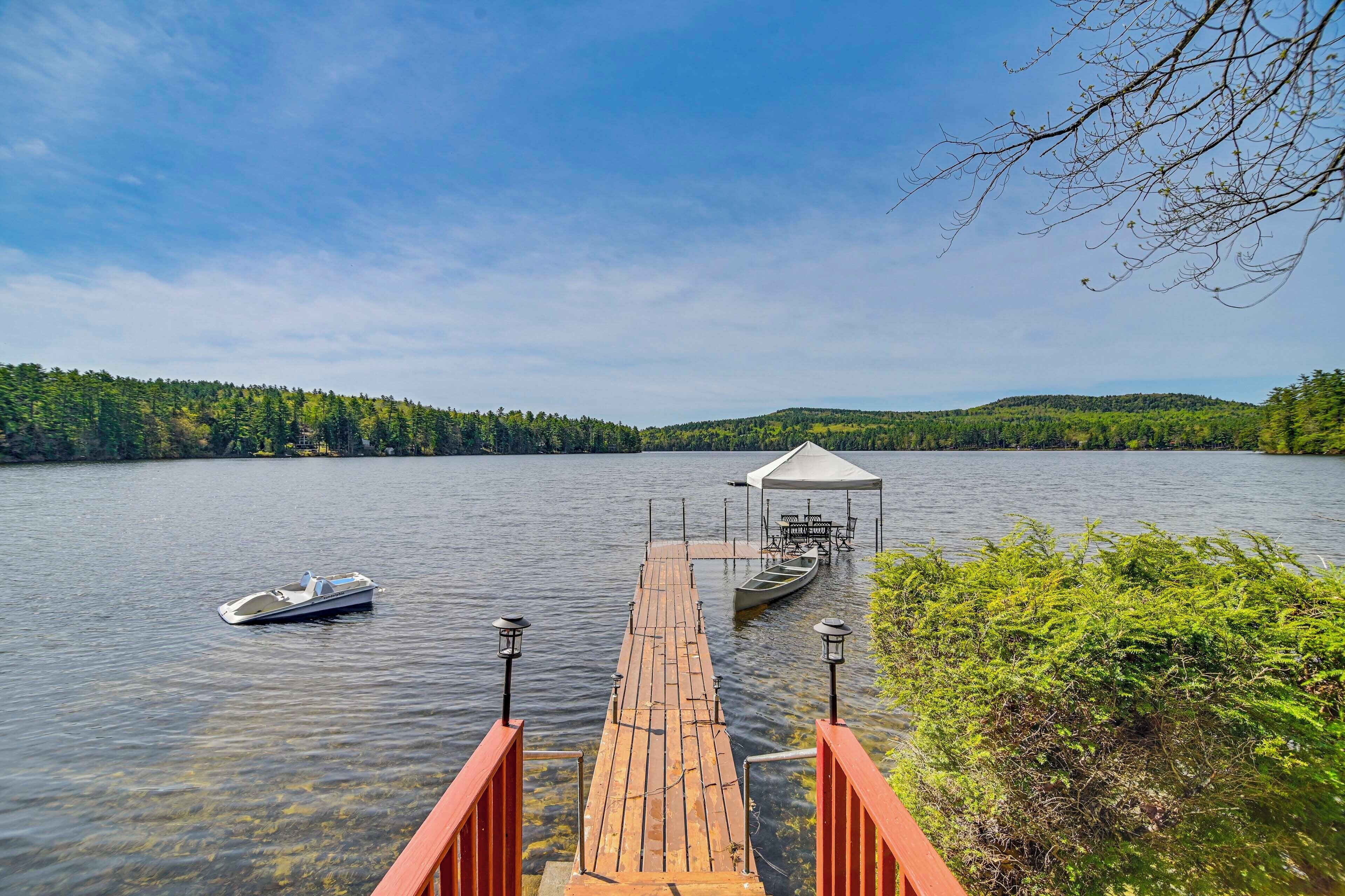 A wooden dock extending into a calm lake, with a small boat and a covered seating area at the end.