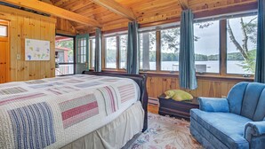 A wooden cabin with a large bed, a blue chair, and a view of a lake.