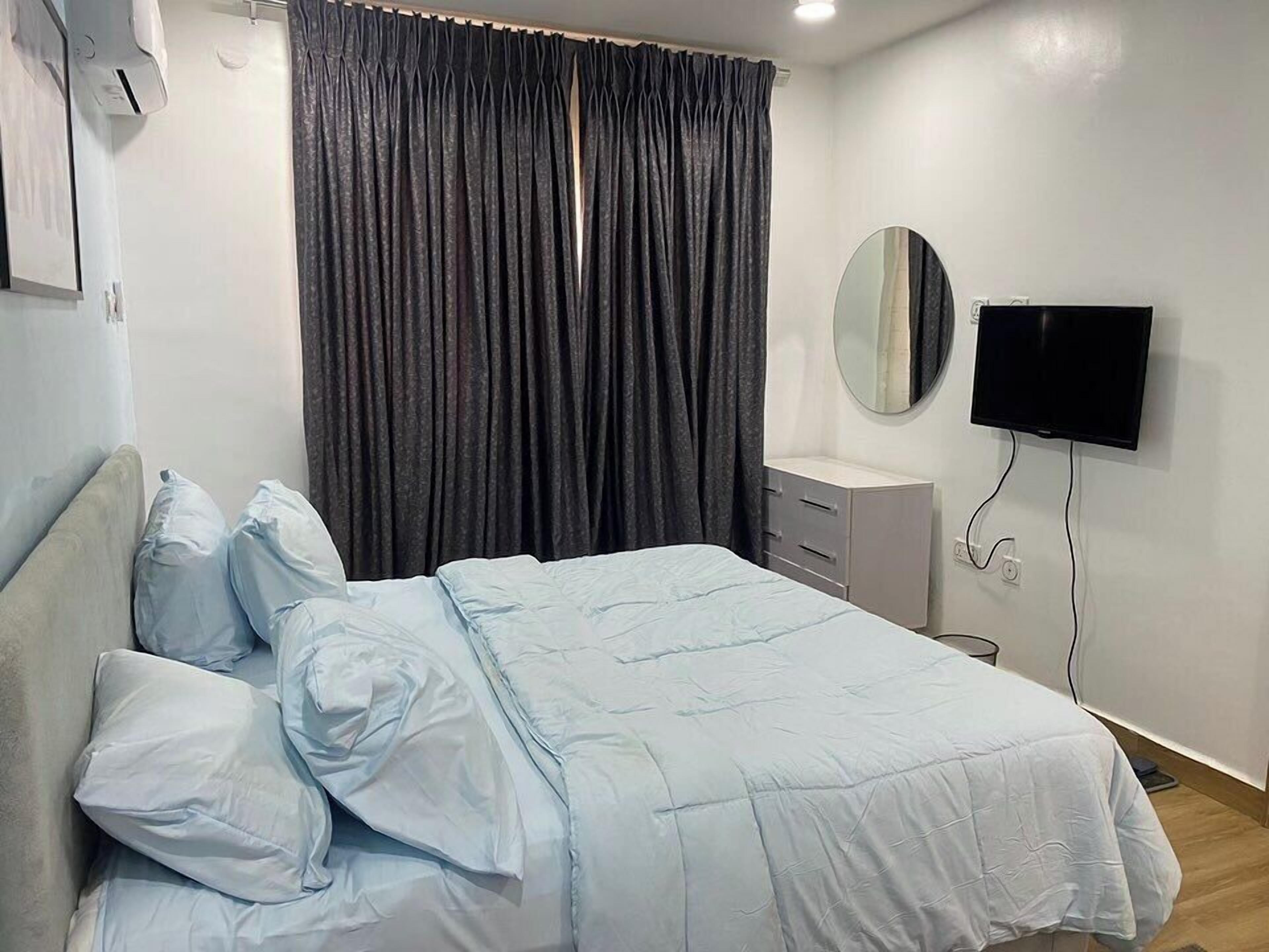 A bedroom with a bed, a bedside table, a mirror, a TV, and a window with curtains.