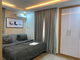 Quarto moderno com cama, roupa de cama cinzenta, uma obra de arte emoldurada, guarda-roupa e janela com cortinas.
