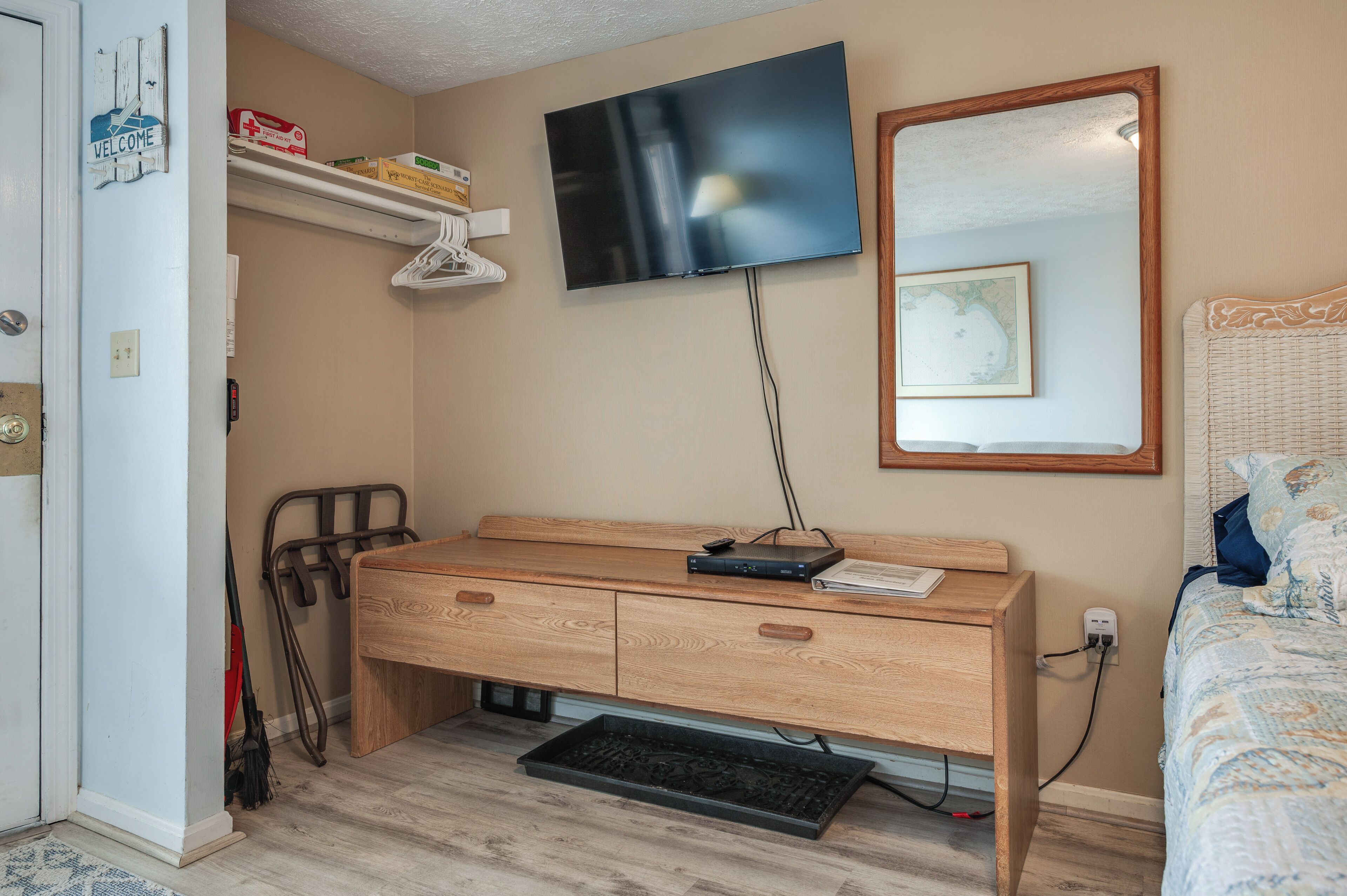 Une chambre avec un bureau en bois, un téléviseur fixé au mur, un lit recouvert d’une couvre-lit à motifs et une petite étagère avec quelques objets.