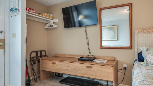 A bedroom with a wooden desk, a TV mounted on the wall, a bed with a patterned bedspread, and a small shelf with items. - Beachfront Unit 17 Fall Escape by the Ocean! (Old Orchard Beach)
