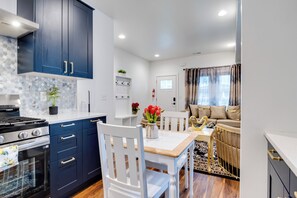 Una cocina moderna con gabinetes azul oscuro, una encimera blanca y una estufa. Hay un comedor con una mesa y sillas blancas, y una sala de estar con un sofá y una ventana.
