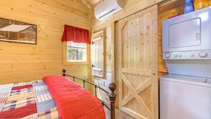 A wooden cabin room with a bed, an air conditioning unit, a washing machine, and a window with a red curtain.