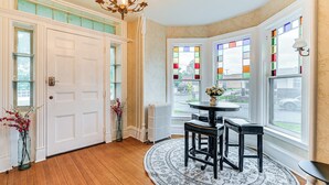 A corner room with a round table, two chairs, a chandelier, a radiator, and stained glass windows.