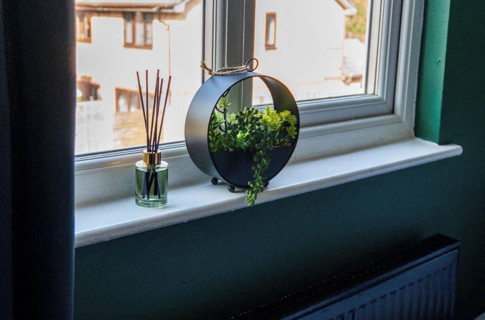 Ein Fensterbrett mit einem runden schwarzen Pflanzgefäß, das grüne Pflanzen und eine Glasflasche mit Schilfhalmen enthält.