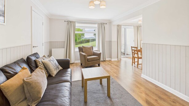 A spacious living room with a leather sofa, a wooden coffee table, and a view of a garden through large windows. - Host Stay The Fairways (Stanley)