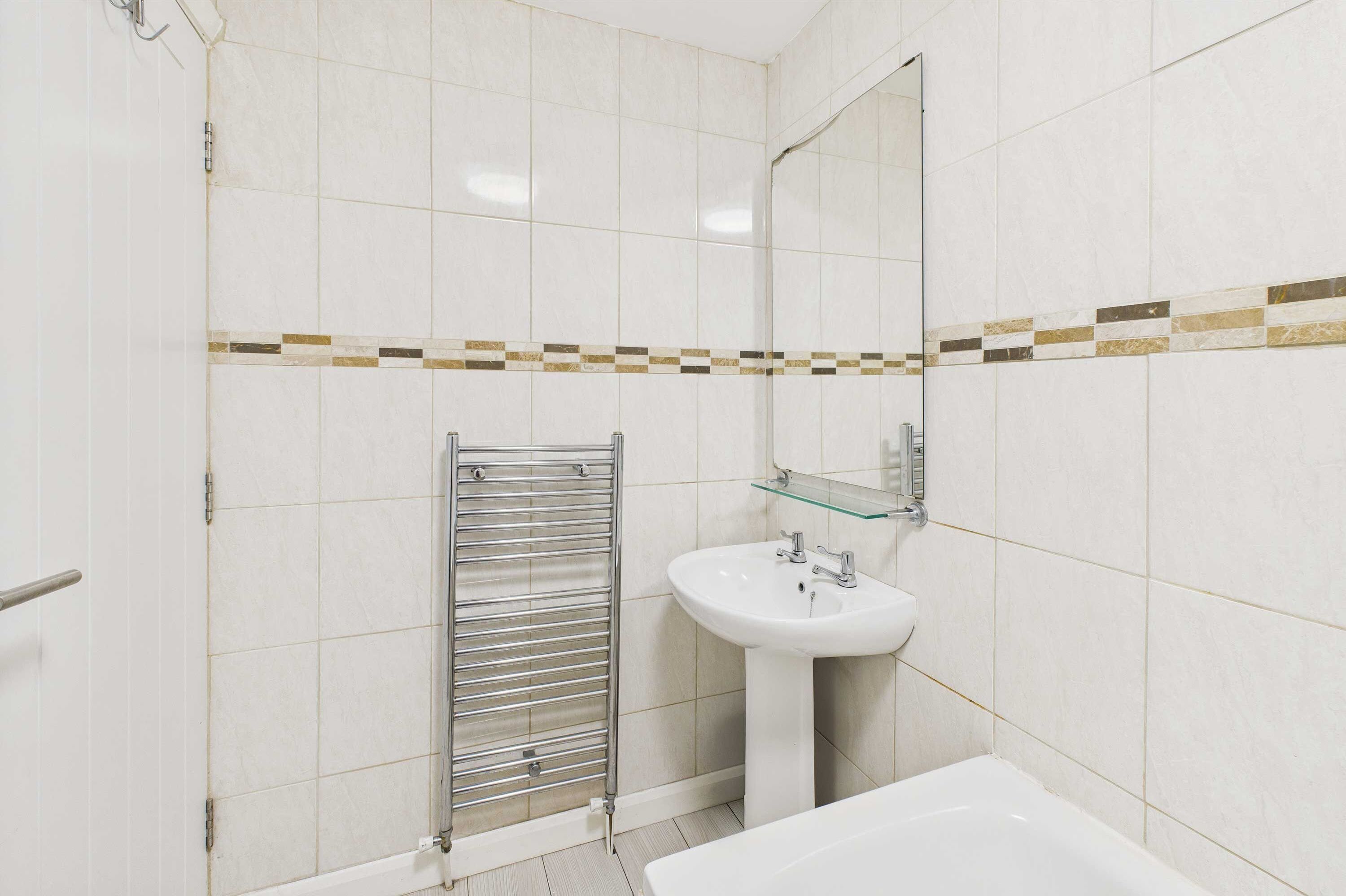 Casa de banho com paredes de azulejos brancos, cabine de duche em vidro, lavatório branco e espelho com bancada de vidro.
