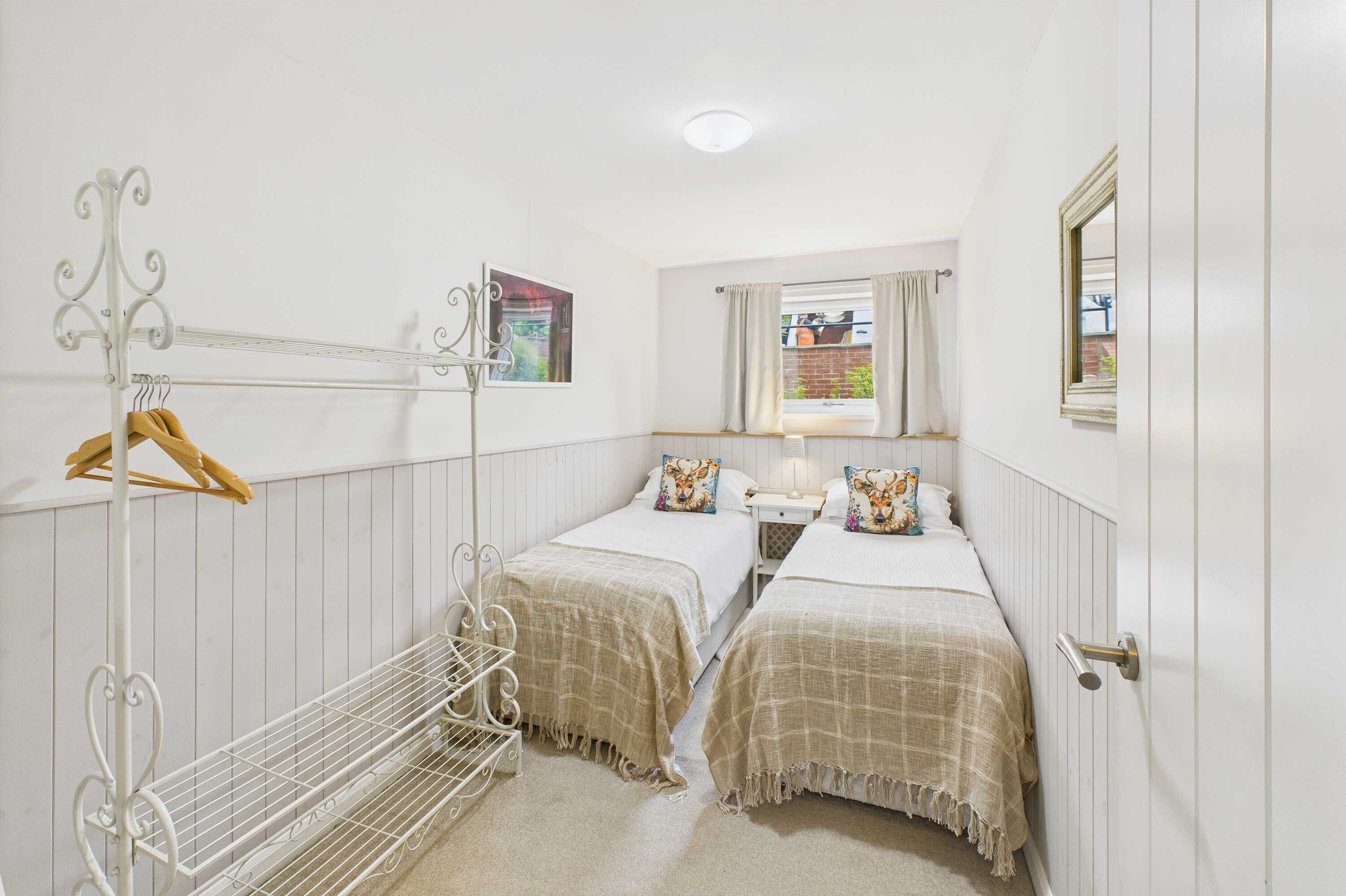 A small, clean room with two beds, a hanging clothes rack, a window with curtains, and a picture on the wall.