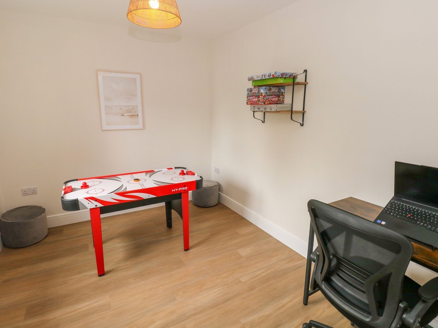 A room with a foosball table, a desk with a laptop, a chair, a shelf with board games, and a hanging lamp.
