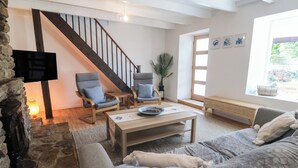 A cozy living room with a stone fireplace, a wooden staircase, a flat-screen TV, two armchairs, a sofa, and a window with artwork. - Candle Croft (Redruth)