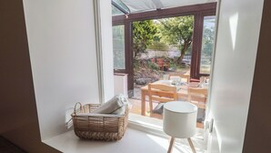 A room with a large window, a wicker basket with cushions, and a white table lamp. - Candle Croft (Redruth)