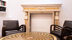 Une pièce comprenant une table ronde ornée d’un motif géométrique, deux fauteuils en cuir foncé et une cheminée agrémentée de moulures décoratives et de bustes.
