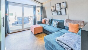 Ein modernes Wohnzimmer mit einem großen Schiebeglasfenster, das auf einen Balkon führt, einem blauen Sofa mit Kissen und Holzboden.