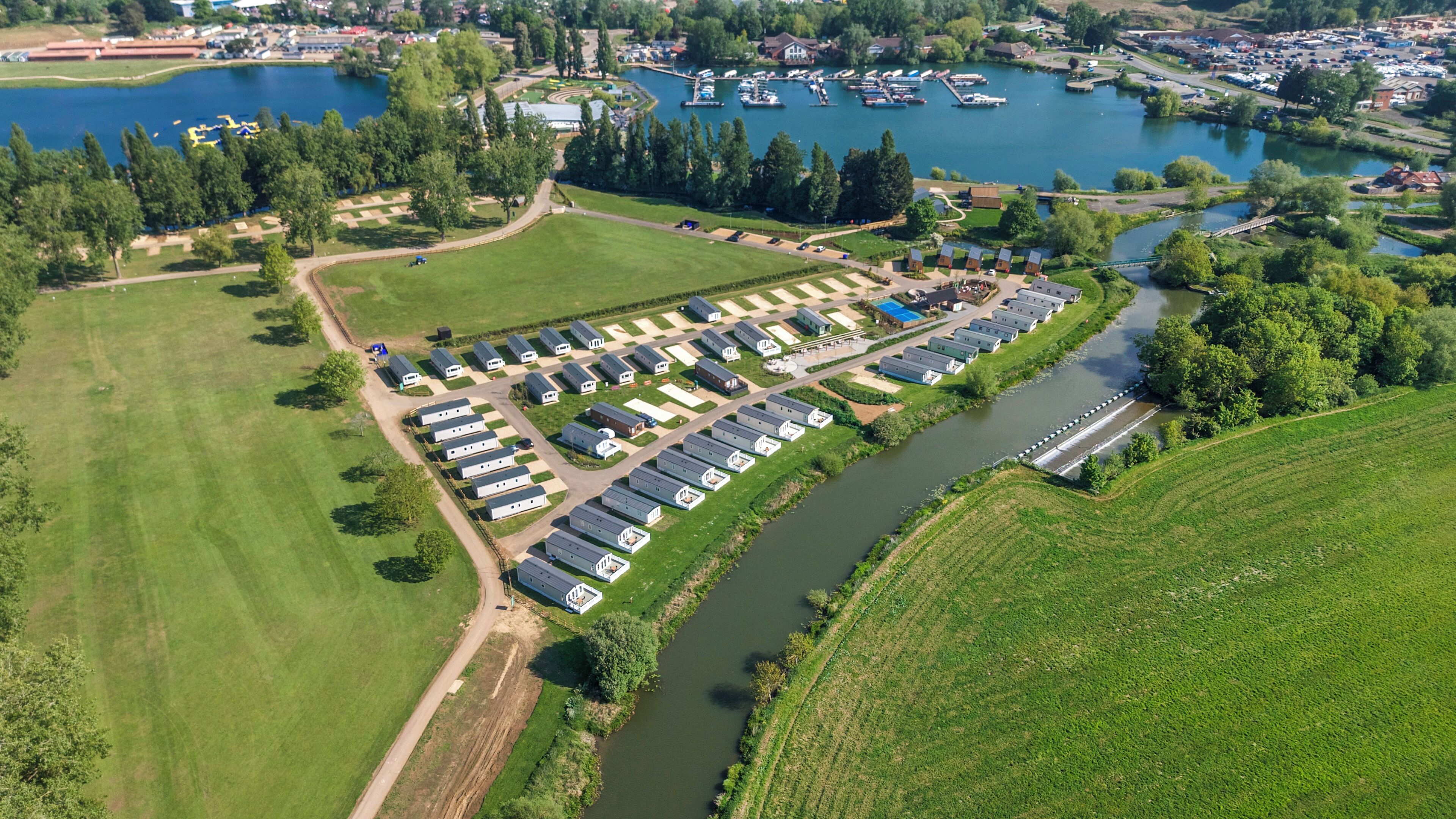 An aerial view of a campground with RVs and mobile homes arranged in rows along a river, surrounded by green fields and a lake with boats.