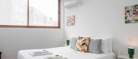 A hotel room with a large window, a bed with neatly folded towels, a wall-mounted air conditioner, and a small bedside table with a lamp.