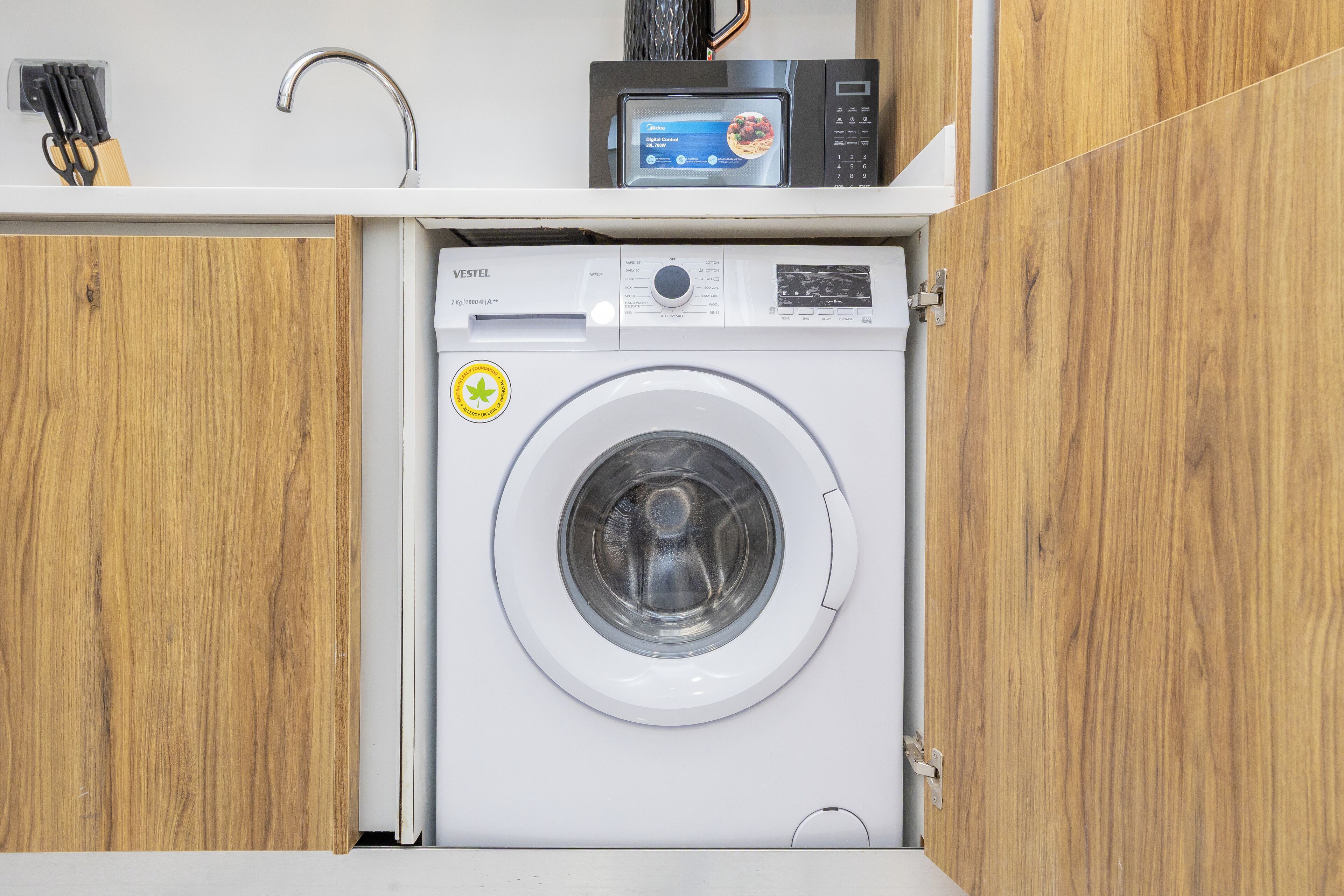 A compact washing machine in a small utility space with wooden cabinets and a microwave above it.