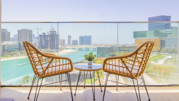 A balcony with two wicker chairs, a small table, and a potted plant. A cityscape with buildings and a bridge is visible in the background.