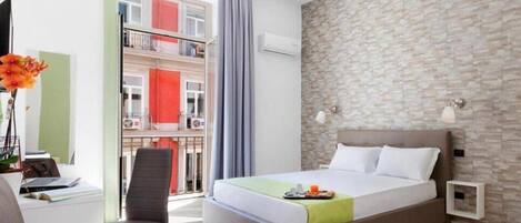 A modern hotel room with a large bed, a desk with a laptop, a chair, and a view of a building with balconies.