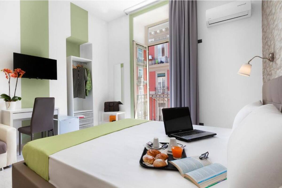 A hotel room with a bed, a desk, a chair, a laptop, a tray with pastries and juice, and a book.