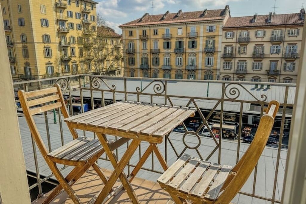 Un balcone con tavolo e due sedie in legno, affaccia su una vista urbana con edifici a più piani.